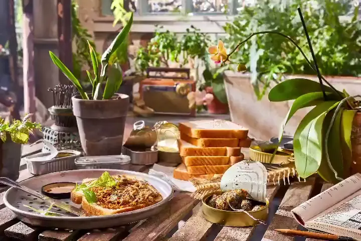 bucolic indoor scene with houseplants and orchids on a wooden table with cans of sardines and stacked slices of bread