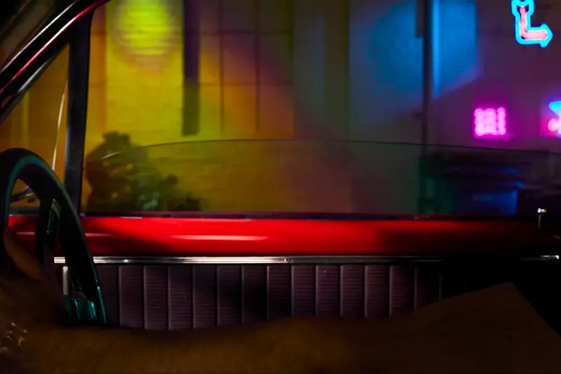 a red vintage car interior on neon lit streeet scene with a hand coming up from lower frame holding a slice of new york style pizza