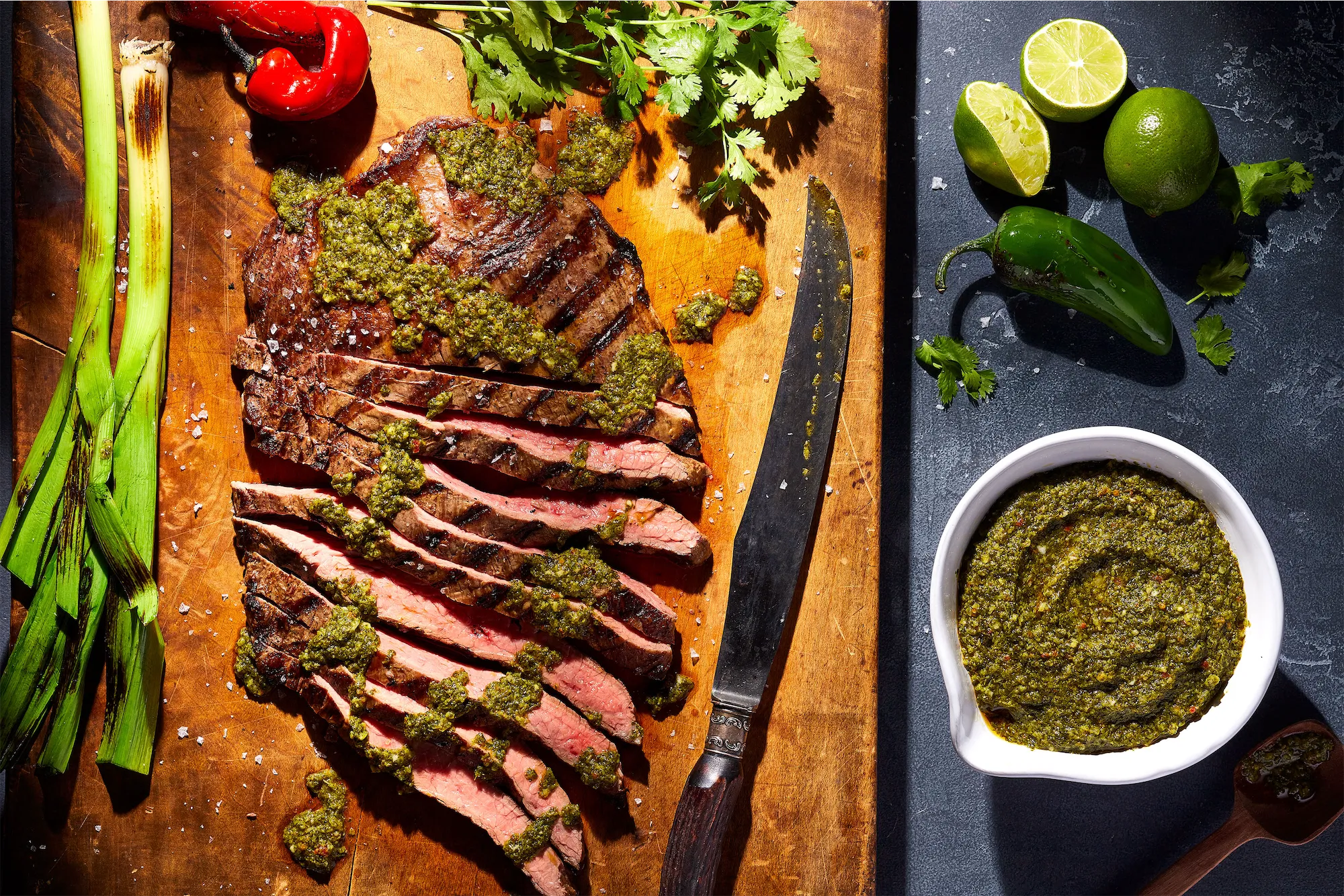 sliced grilled steak on a wooden board with chimichurri salsa limes jalapenos and grilled green onions