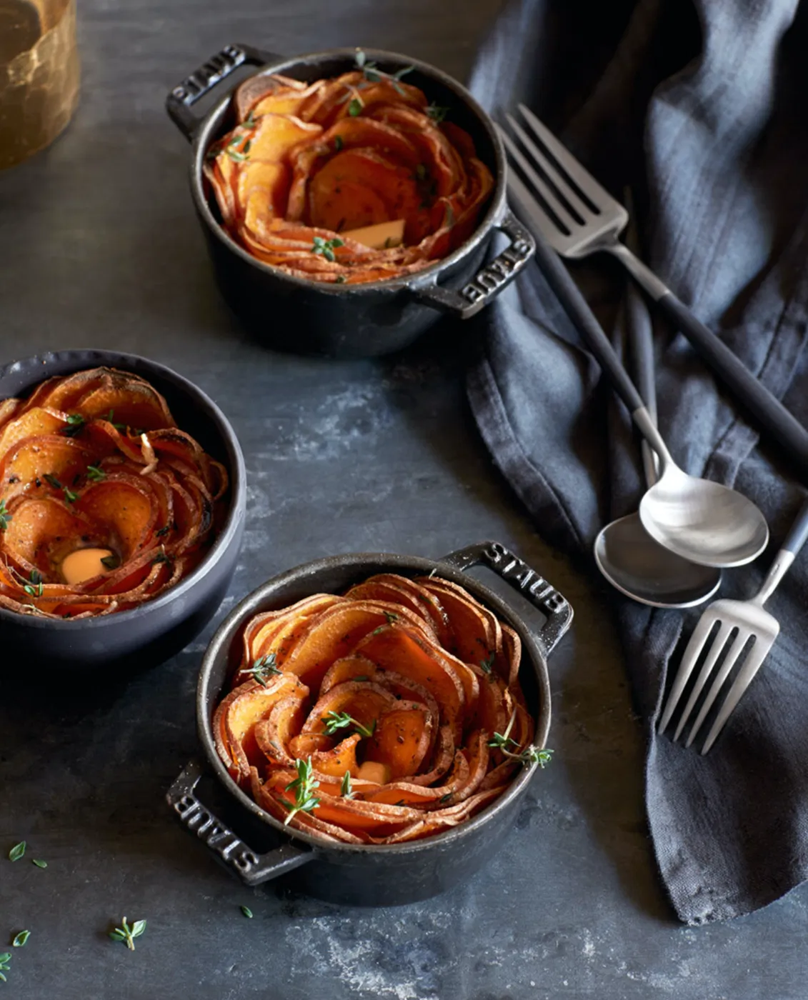 three mini staub bakers with sweet potato layers on grey background and contemporary stainless brushed flatware