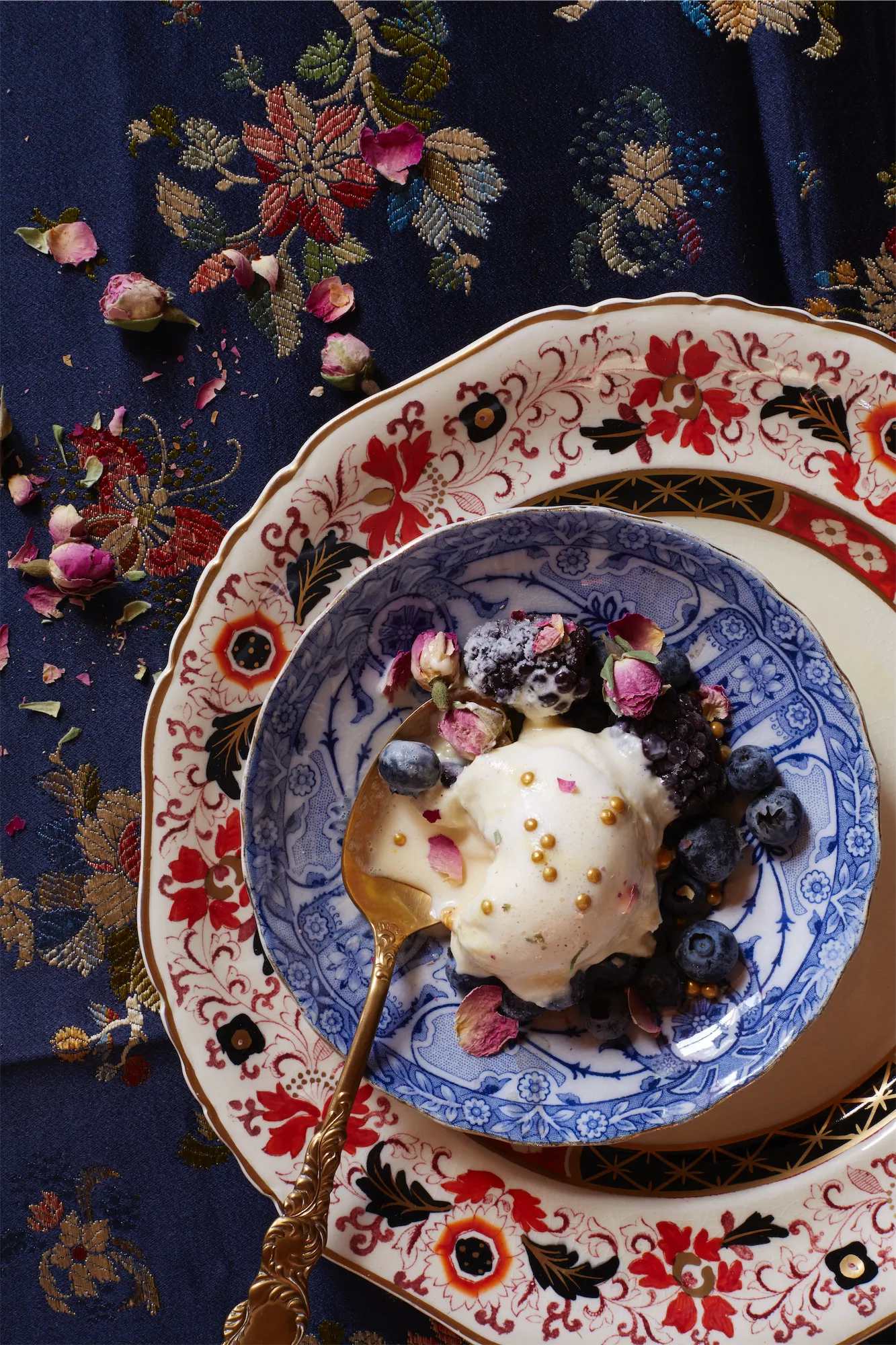 scoop of ice cream with blueberries and rose petals on layered antique plates set on vintage silk embroidered fabric