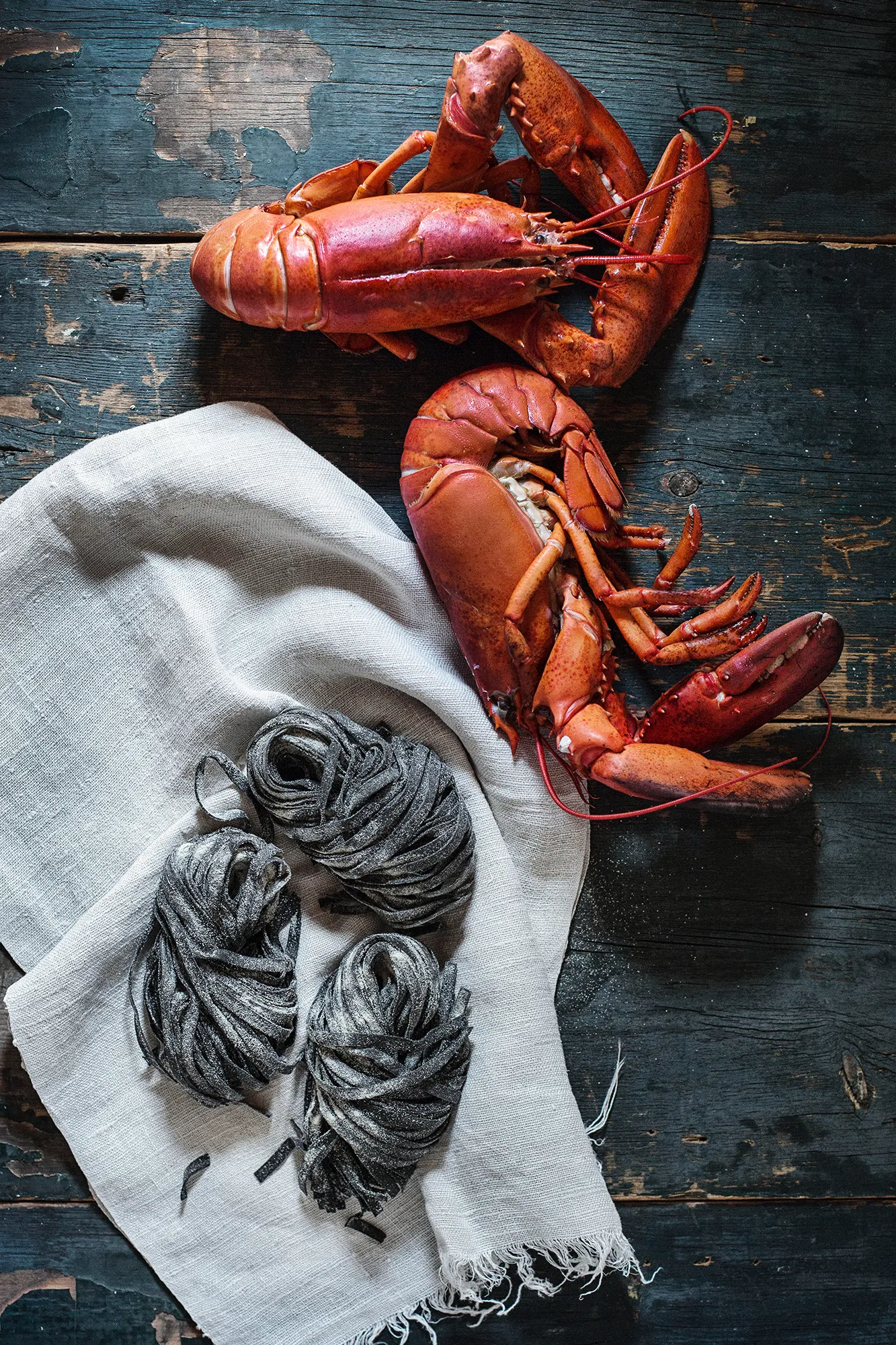 a pair of cooked lobsters on a rustic blue wood surface with a white cloth holding three bundles of squid ink pasta