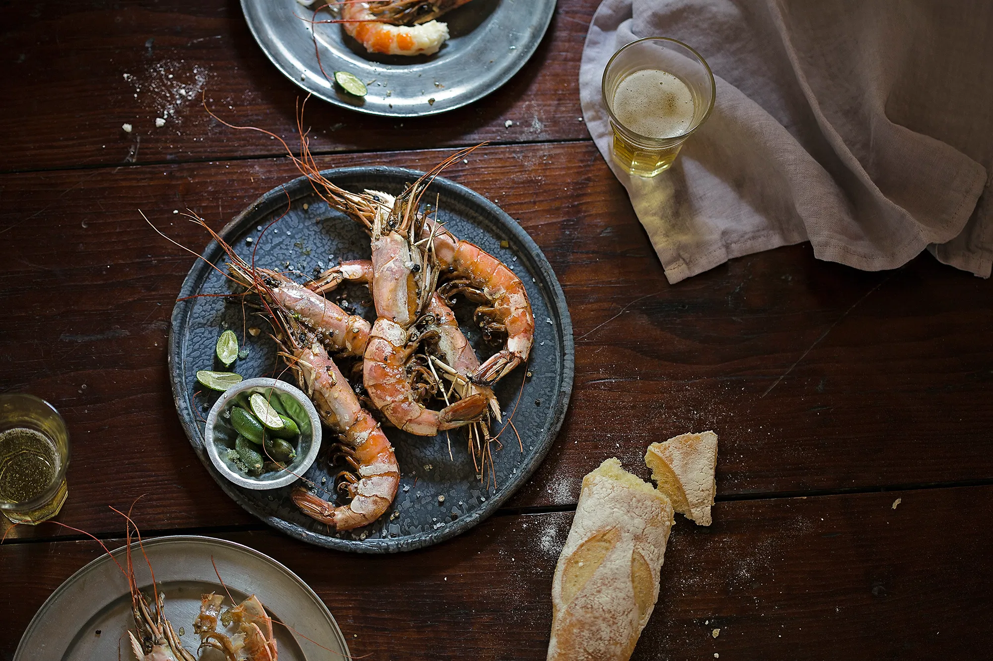 a plate of cooked shrimp with torn baguette pewter plates and glasses of beer on rustic wood table