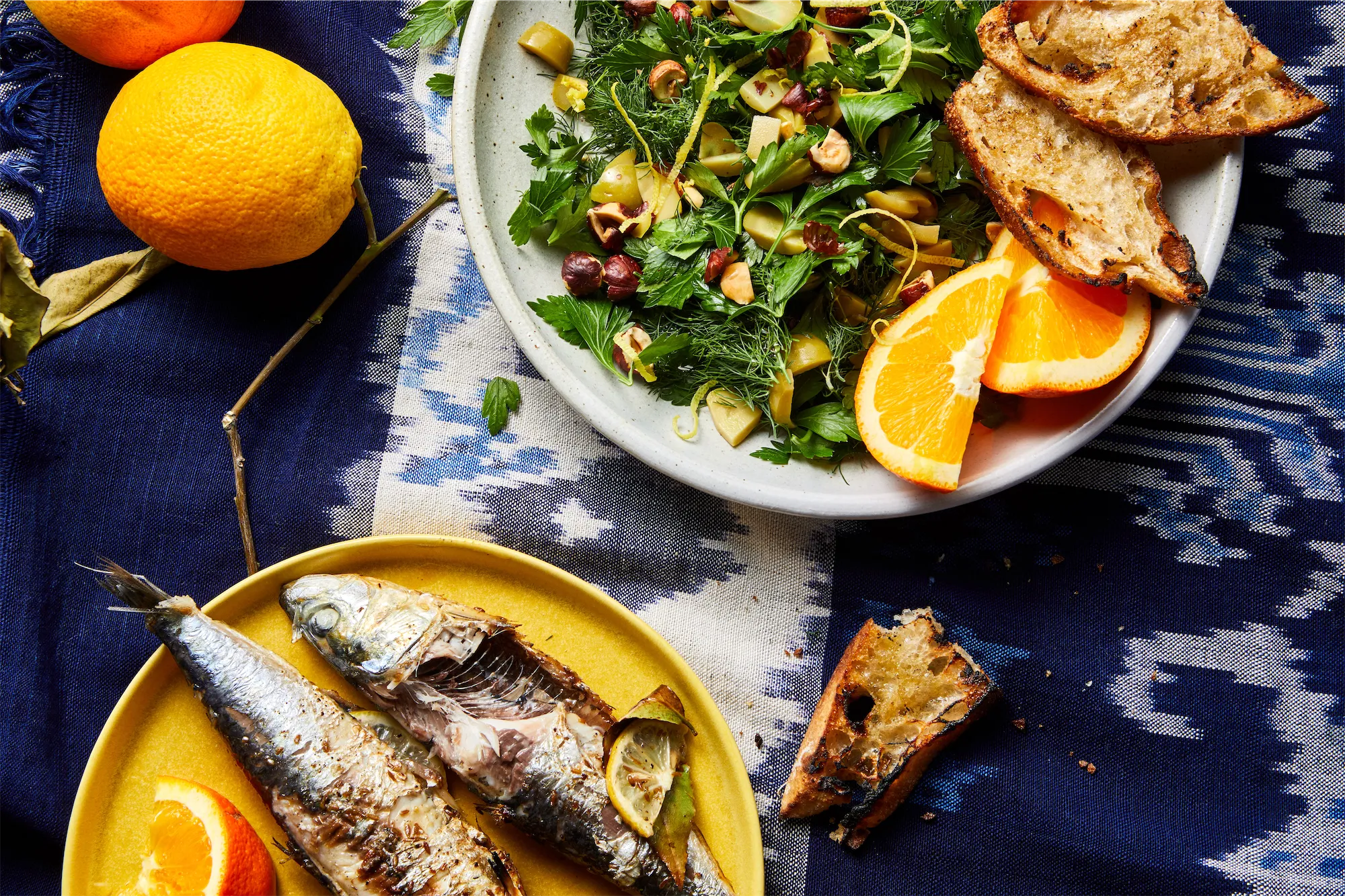 fresh grilled sardines on yellow plate with olive and hazelnut green salad oranges and crusty bread on a blue and white tablecloth