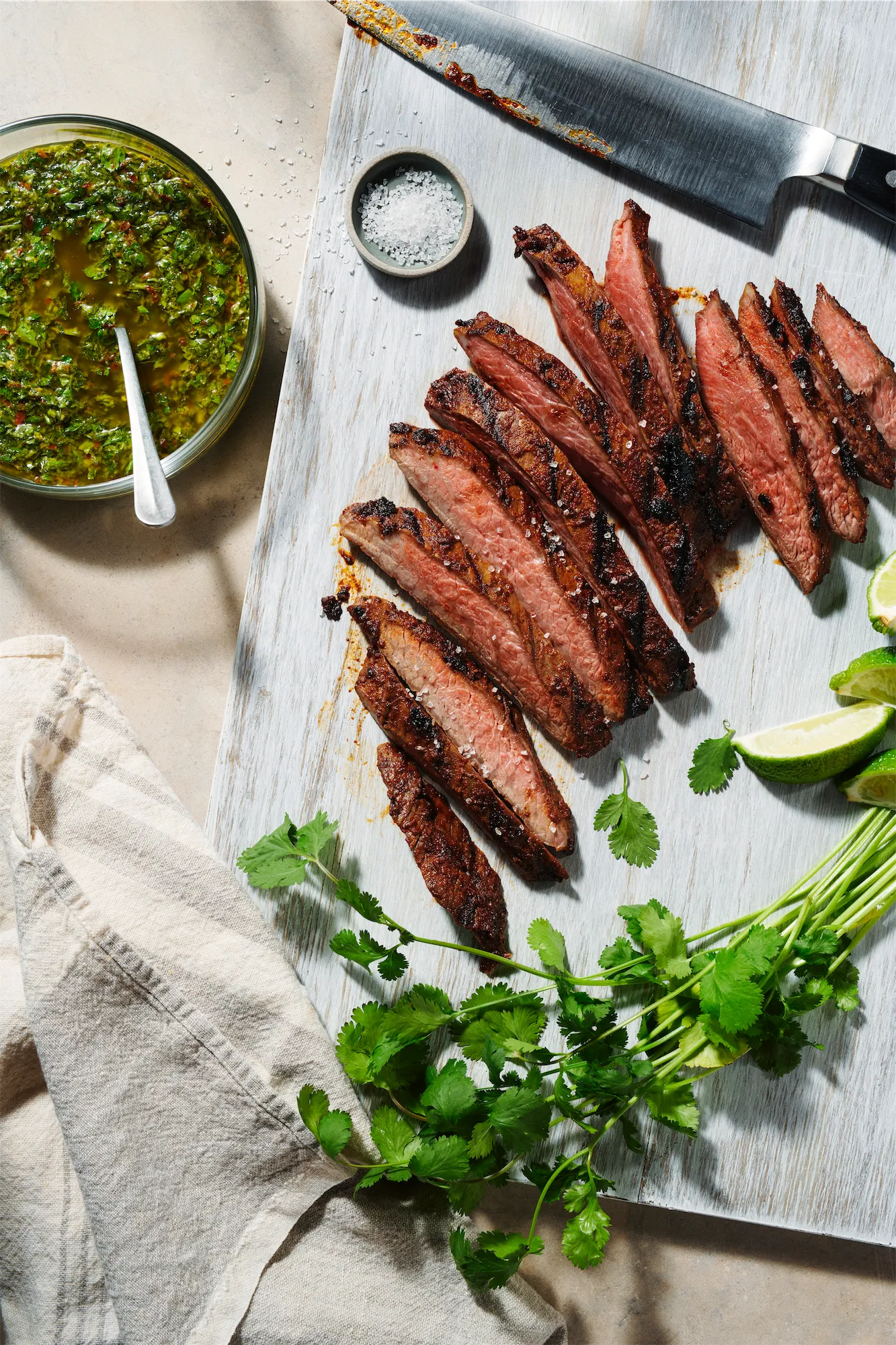 sliced rare staek on a white cutting board with coarse morton salt and a green salsa