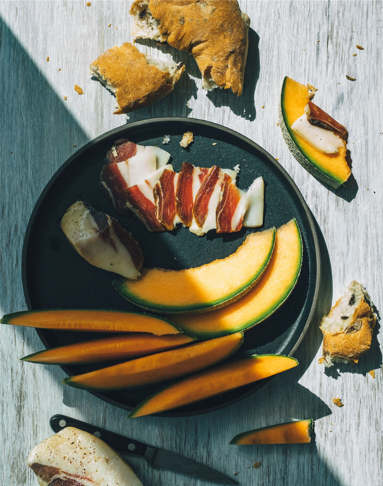 plate of melon and proscuitto with crusty bread and long shadow