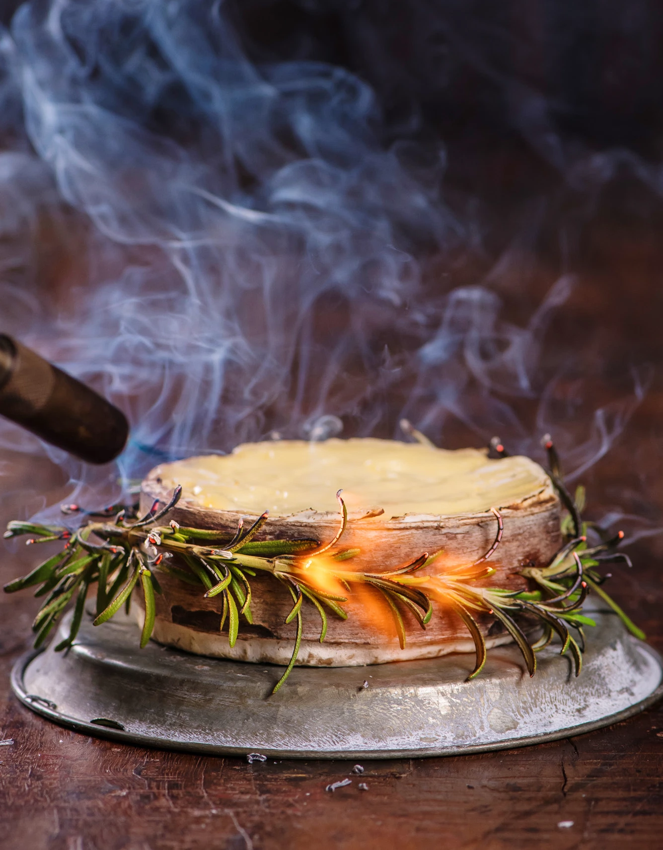 Harbison cheese wrapped in rosemary and smoking with a blow torch flame