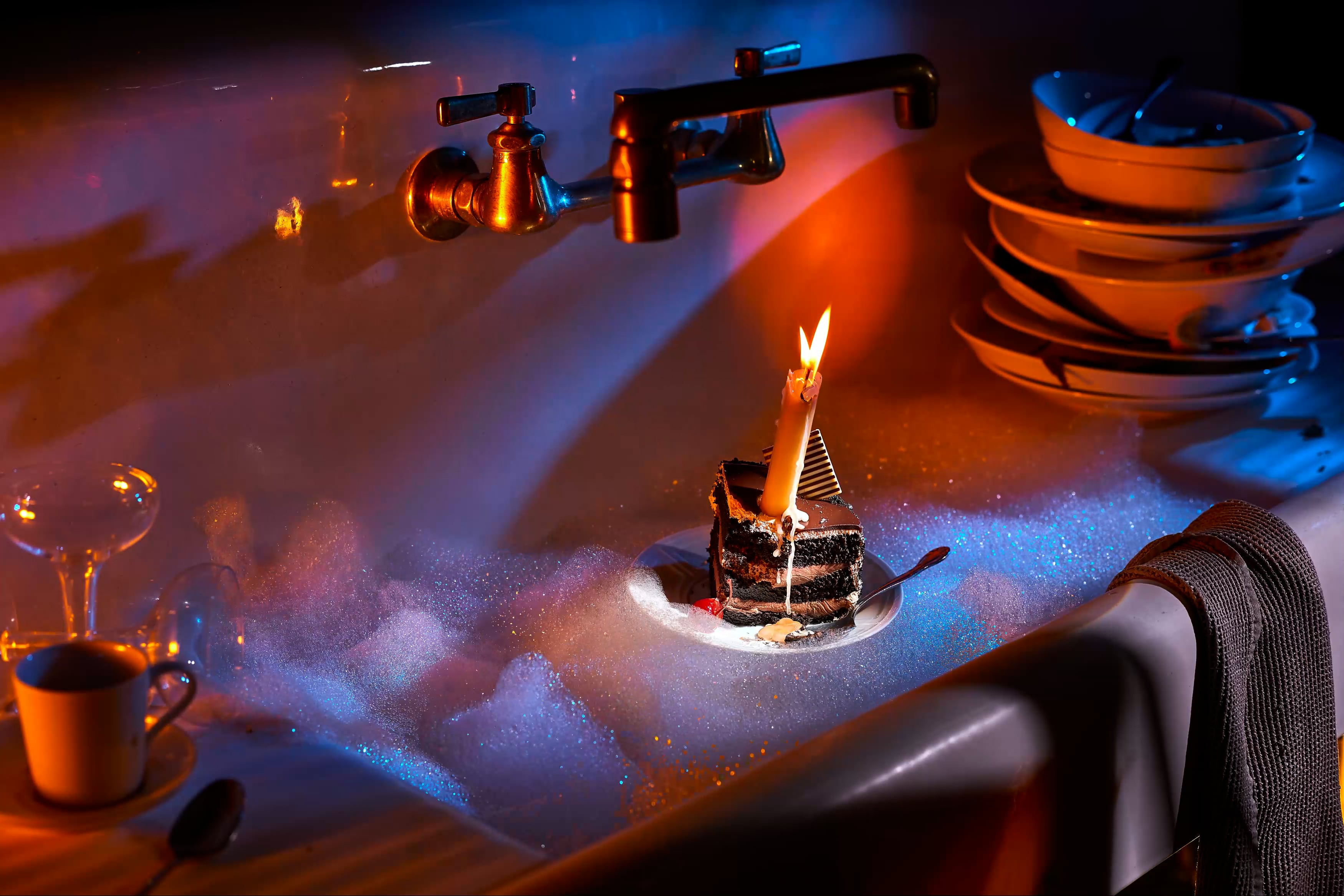 slice of celebration cake with a flickering candle floats in a sudsy vintage sink with piles of dirty dishes