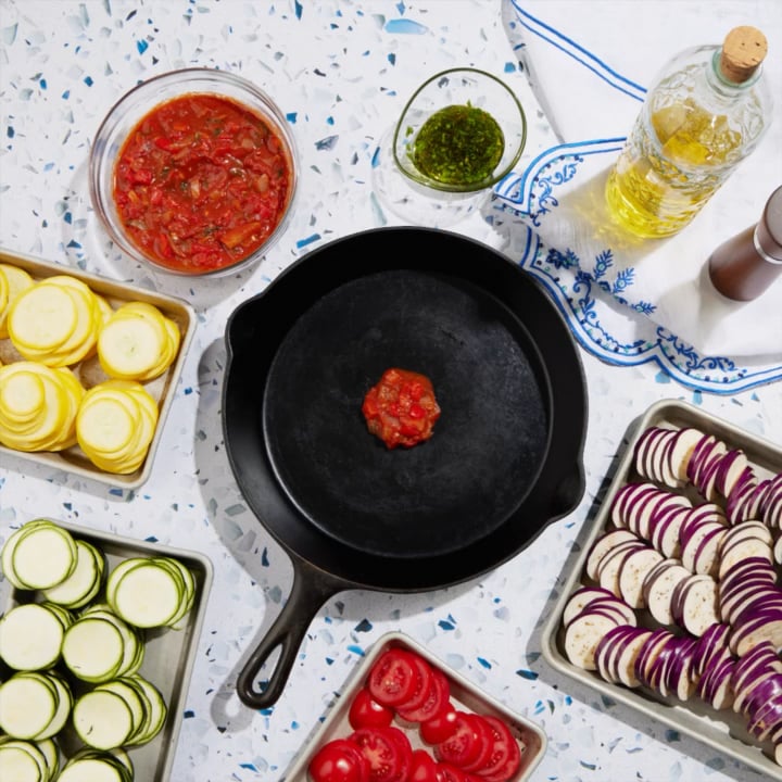 step by step building zucchini eggplant yellow sqash tomato in a cast iron skillet making ratatouille on blue terrazzo surface