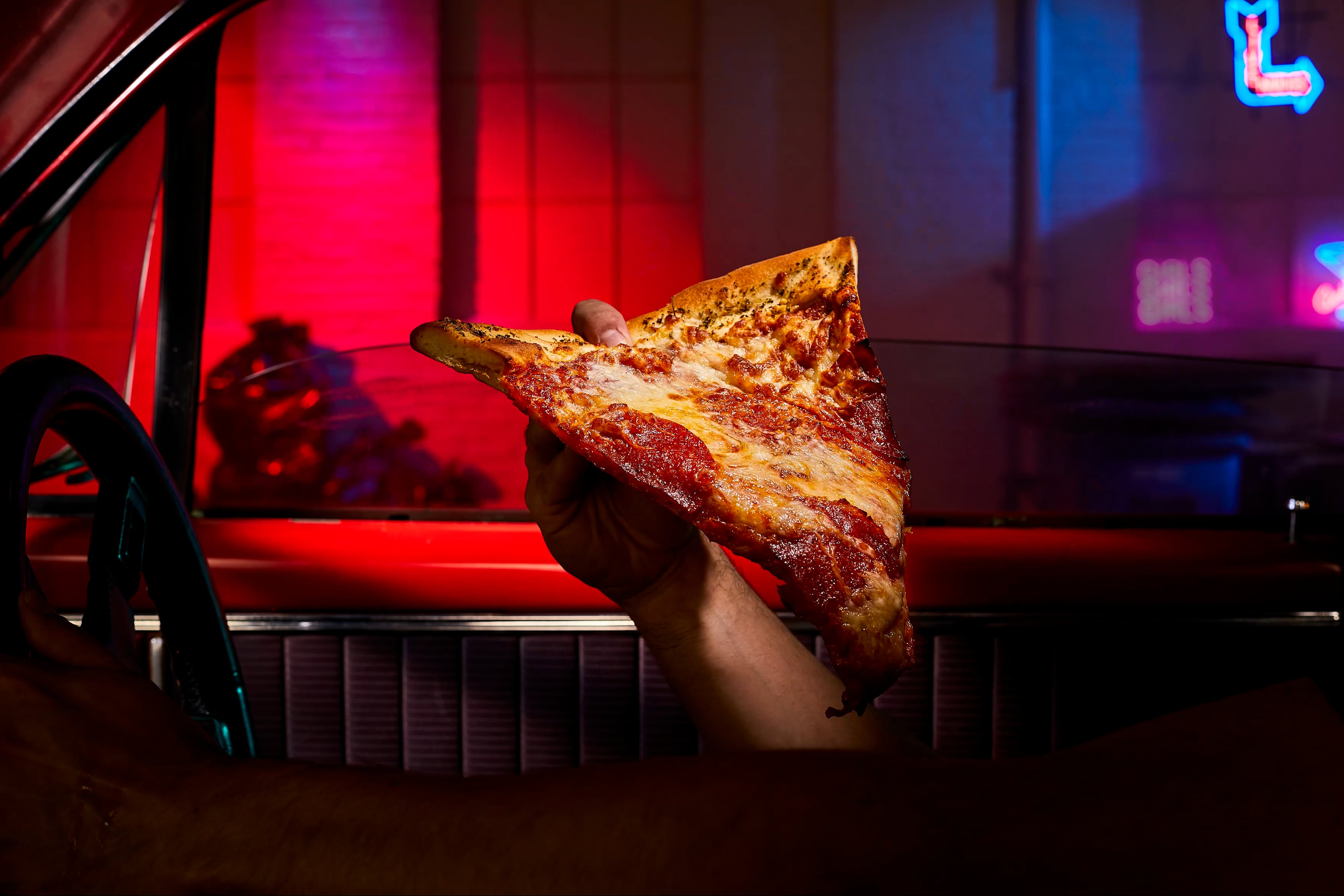 a red vintage car interior on neon lit streeet scene with a hand coming up from lower frame holding a slice of new york style pizza