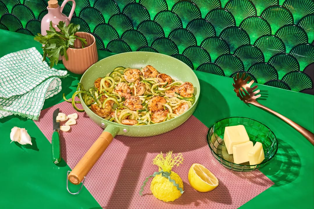 Pescanova brand shrimp and zucchini noodles in a ceramic pan on vibrant green backround and pink placemat with fresh lemon parsley and cubes of butter