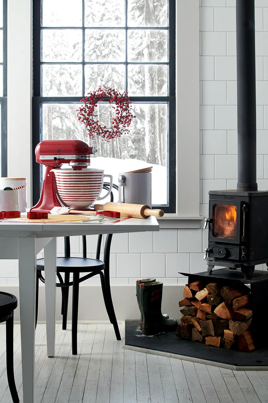 Black and white kitchen scene with hobbit woodburning stove and red kitchenaid standmixer on white table black chairs wood stacks wellies white wood foor crate and barrel winter scene