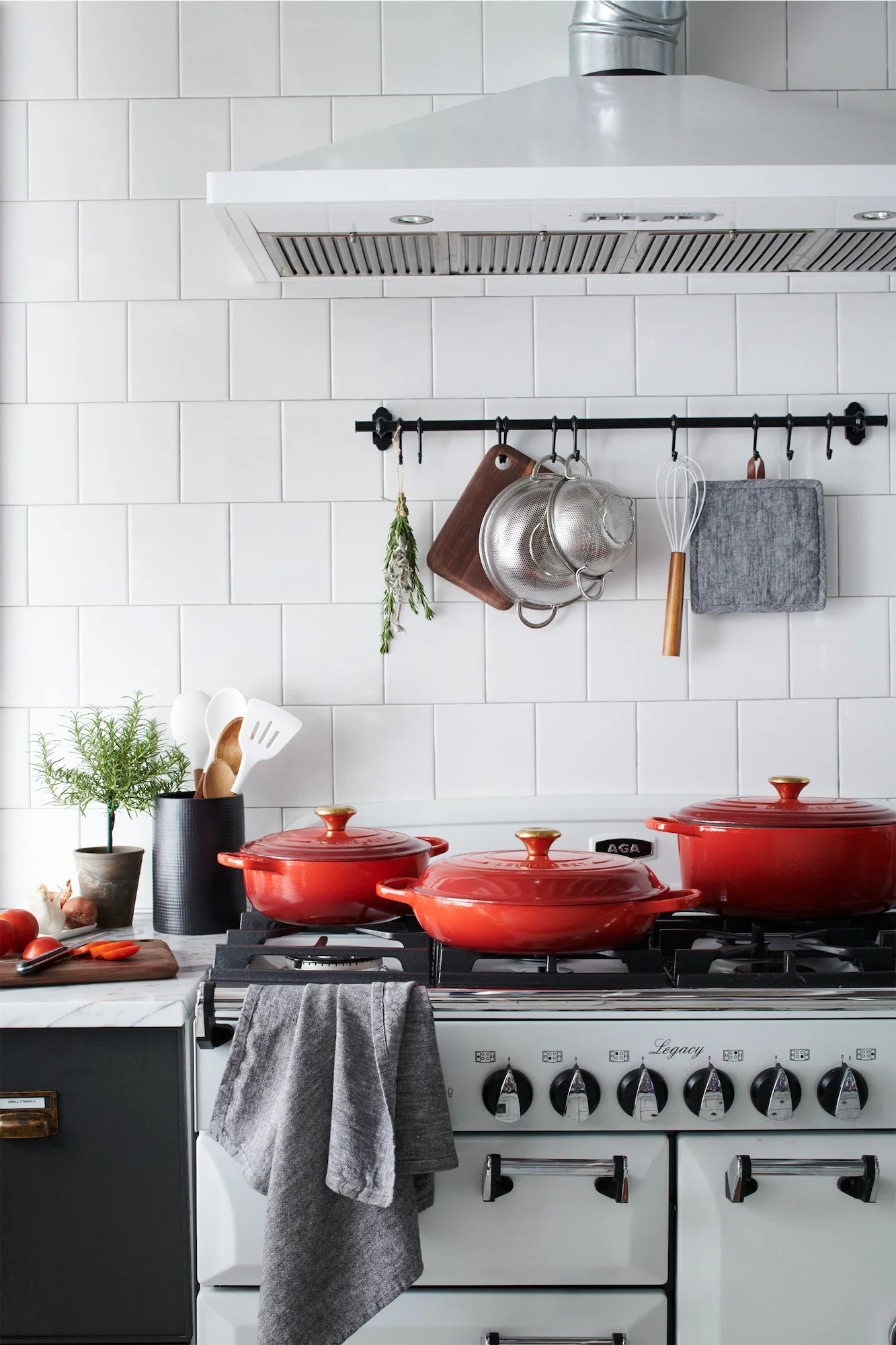 Crate and barrel red le crueset pots and pans on a white AGA in a white tiled kitchen with potted rosemary and hanging utensils tasteful