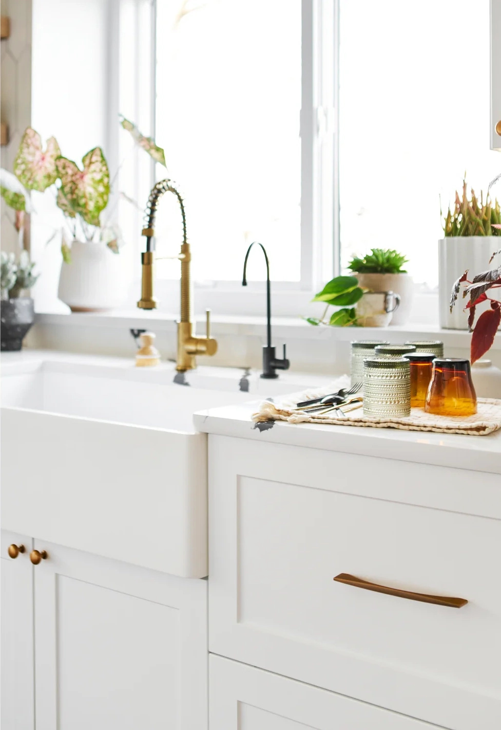 Tasteful white kitchen scene at sink with cabinets brass hardware handles and plants on the windowsill