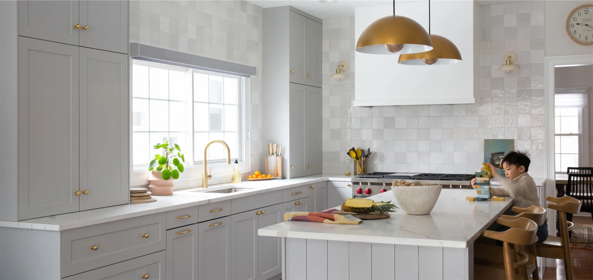SI7 tasteful white kitchen with a large island at which sits a young boy and the signs of preparing a meal gold domed pendants hang above and a large window over the sink