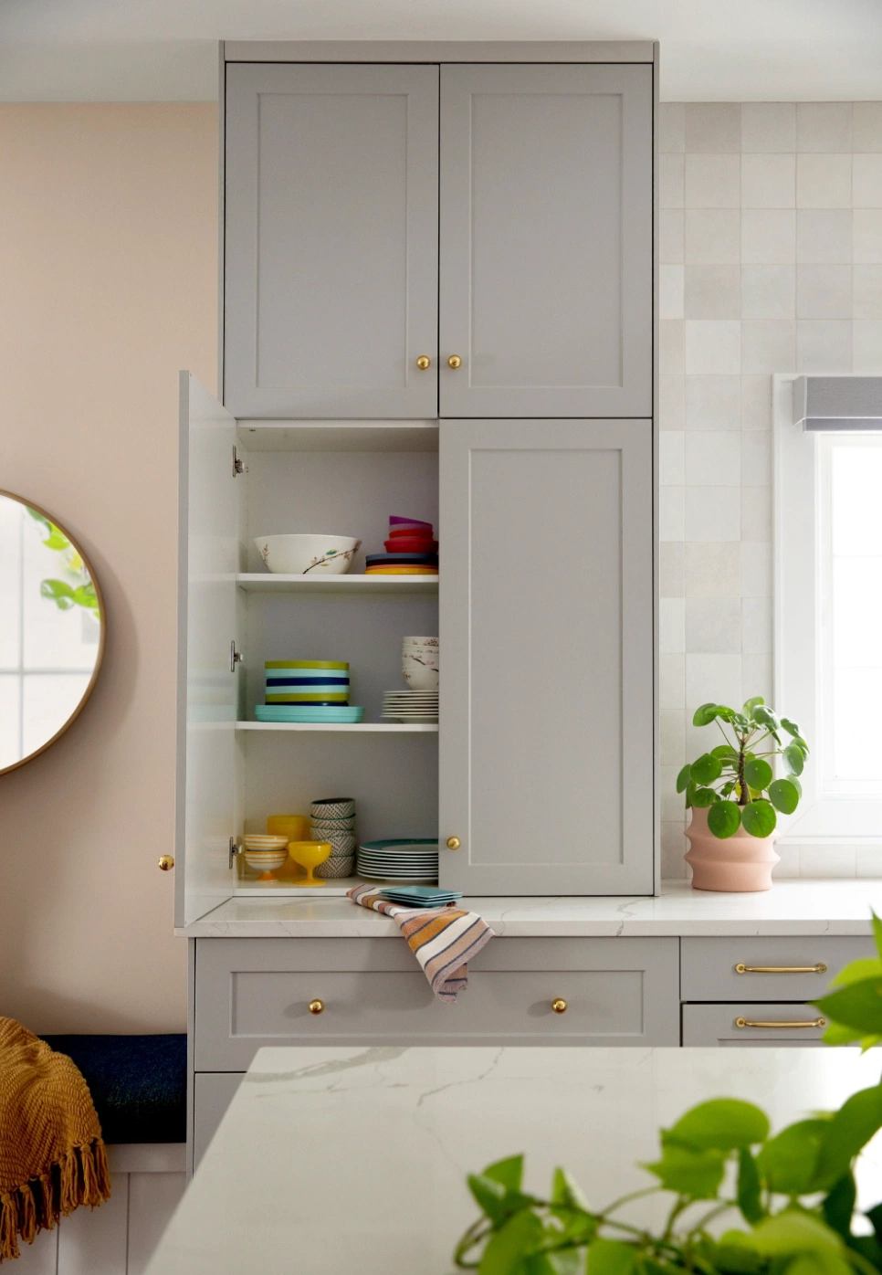 contemporary tasteful kitchen cabinet in pale grey with gold hardware stacks of plates and bowls greenery by the window